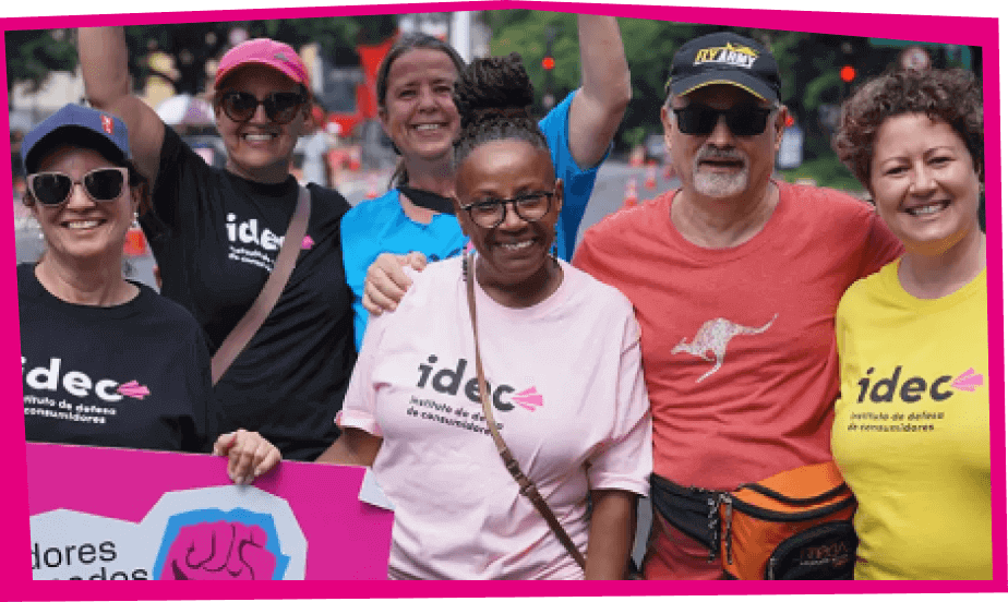 Foto de um grupo de seis pessoas sorrindo e abraçadas, todas vestindo camisetas do IDEC. As camisetas têm várias cores, incluindo preto, rosa e amarelo. Eles estão participando de um evento ao ar livre